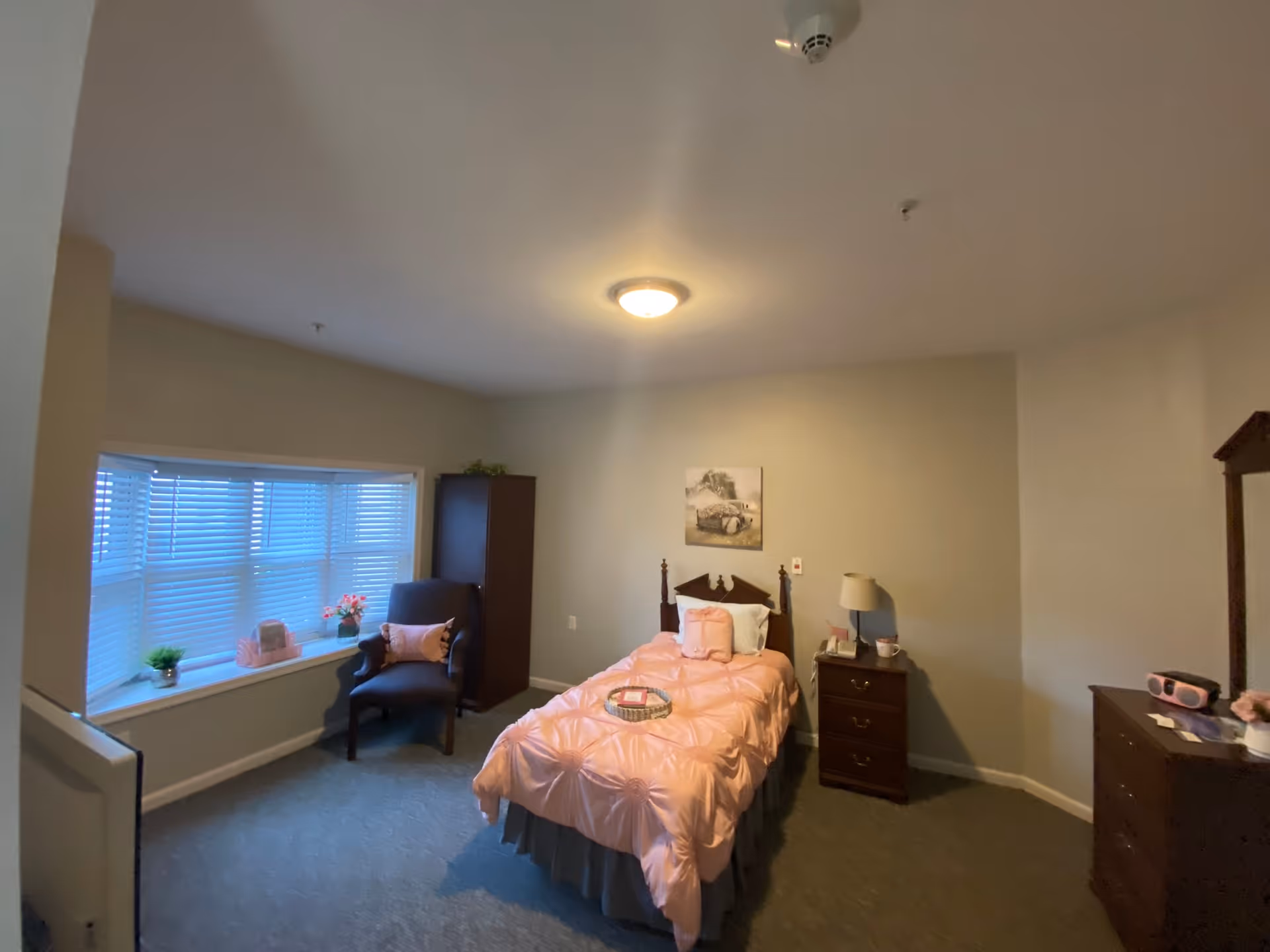 A cozy bedroom in a senior living facility with a single bed covered in a pink comforter and matching pillows. The room has a window with blinds, a dark armchair with a pink pillow, a tall dark cabinet, a nightstand with a lamp and phone, and a dresser with a mirror and small items on top. A framed picture hangs on the wall above the bed.