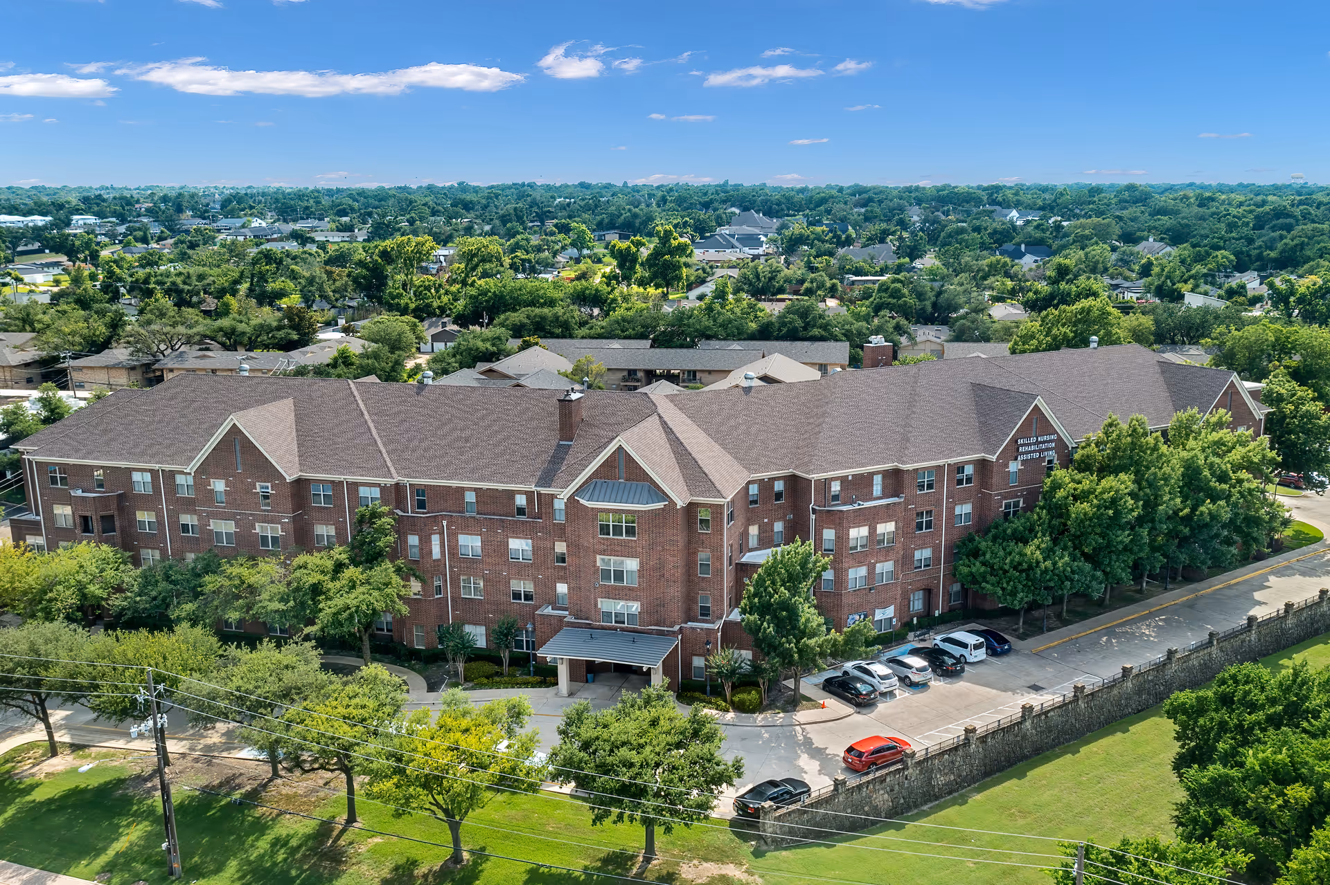 Aerial view of a large, multi-story brick senior living facility surrounded by trees and greenery under a clear blue sky. Several cars are parked in front of the building along a driveway.