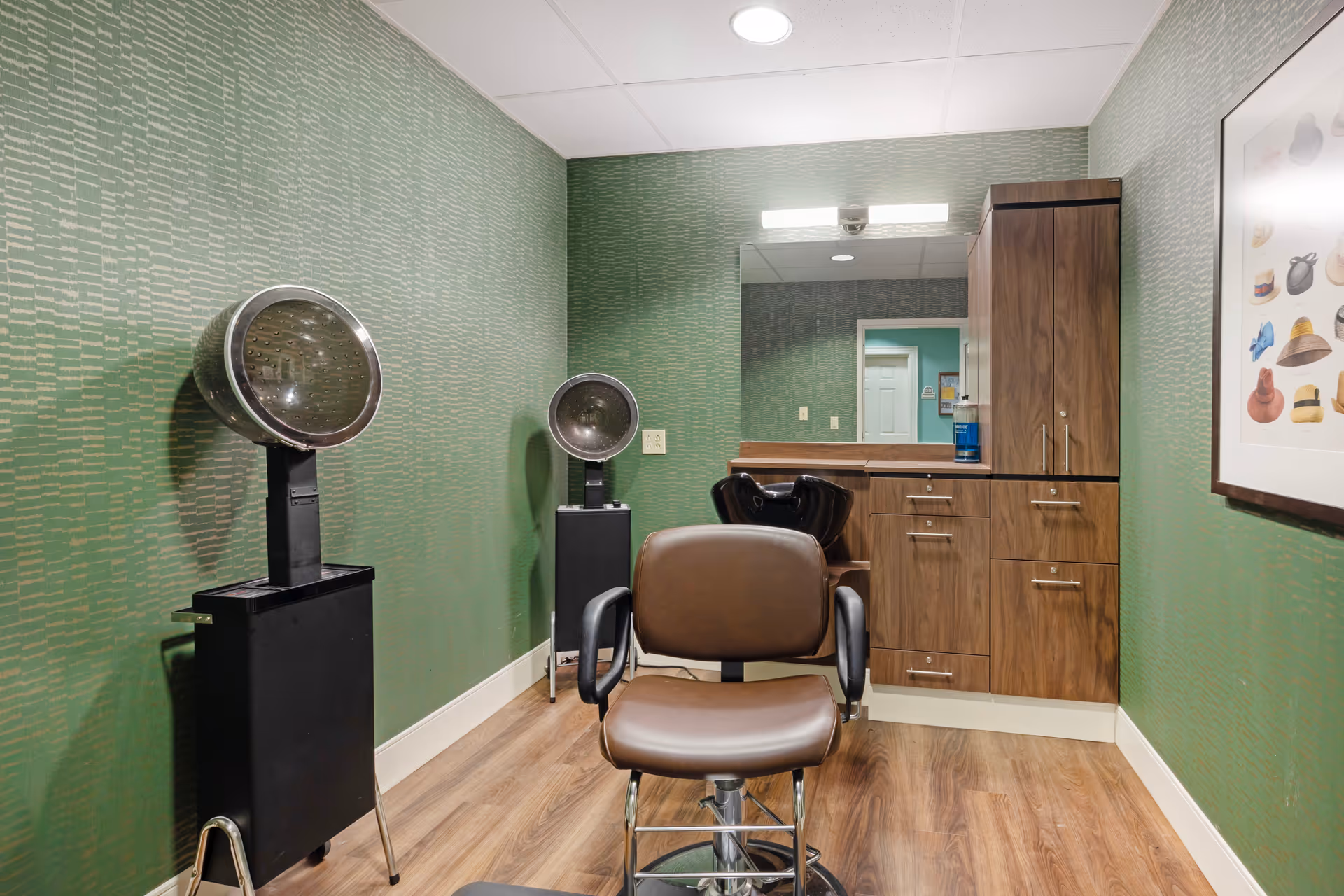 Small salon room with a brown styling chair, hooded hairdryers, a mirror and wooden cabinets against green wallpaper.