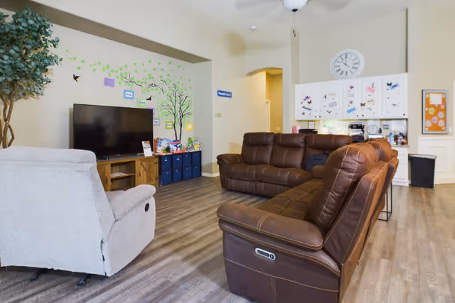 Bright common room with brown leather sofas and a recliner facing a TV, wall decals, storage cubes, and cabinets under a large clock.