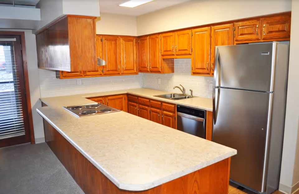 A kitchen with wooden cabinets, a beige countertop, a stainless steel refrigerator, a built-in stovetop, a dishwasher, and a sink. There is a door with blinds on the left side.