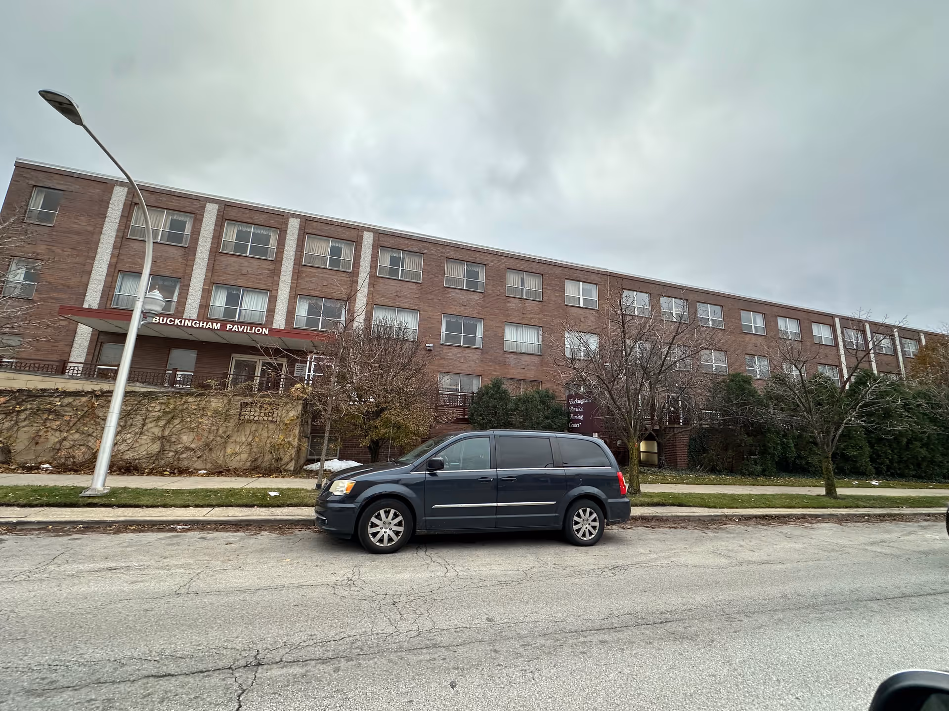 Exterior view of Buckingham Pavilion Nursing and Rehabilitation, a three-story brick building with multiple windows and a red awning over the entrance. A dark-colored minivan is parked on the street in front of the building, and leafless trees and shrubs are visible along the sidewalk. The sky is overcast.