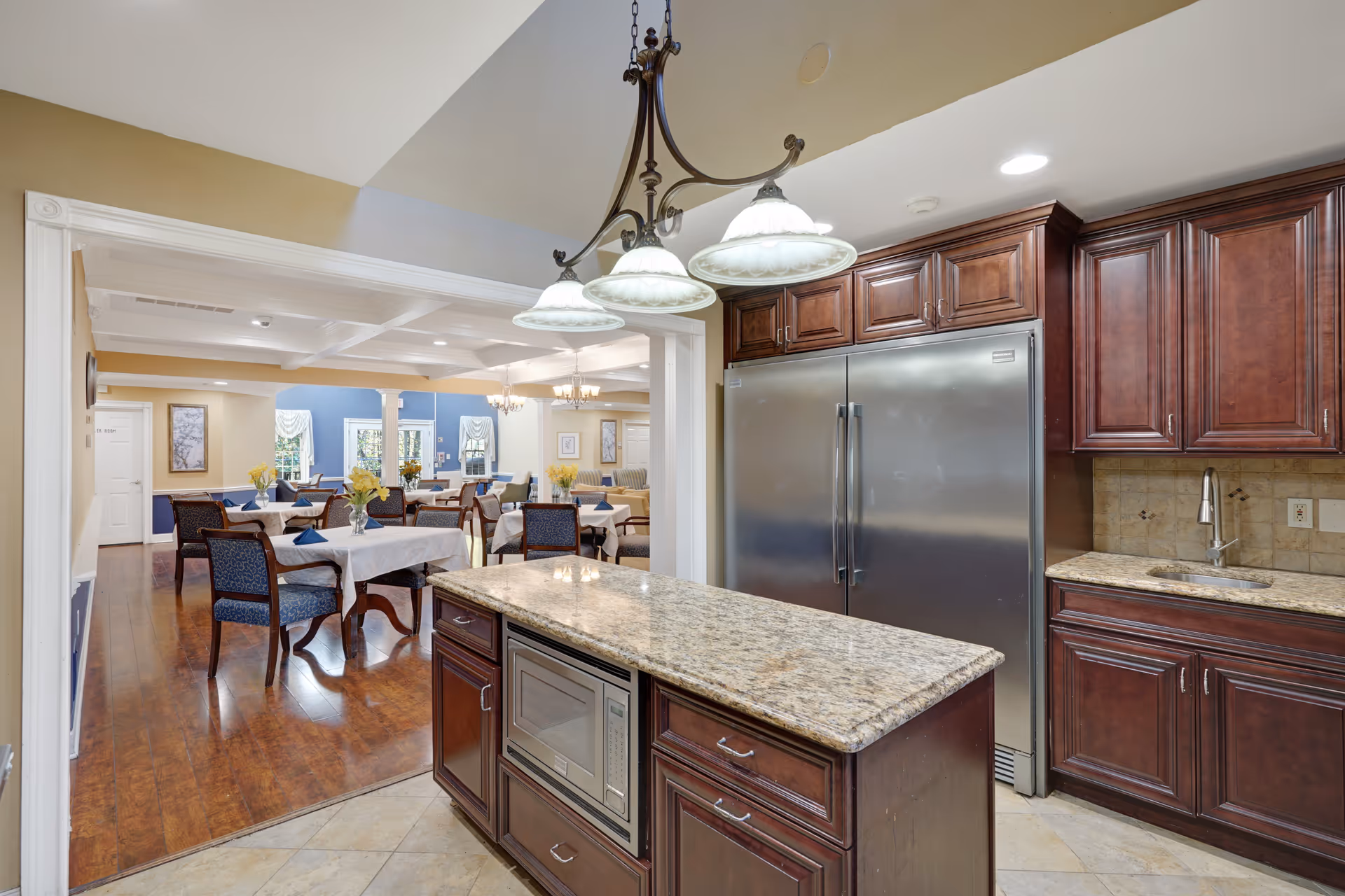 View of a kitchen area with dark wood cabinets, a large stainless steel refrigerator, granite countertops, and an island with a built-in microwave. Beyond the kitchen is a dining room with multiple tables covered with white tablecloths, each set with chairs and small flower arrangements. The dining room has wooden flooring and blue accent walls with windows and framed artwork.