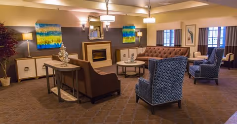 A cozy living room area with a brown tufted sofa, two blue patterned armchairs, a round coffee table, and a fireplace with a mirror above it. The walls are decorated with colorful abstract paintings and there are several lamps providing warm lighting.