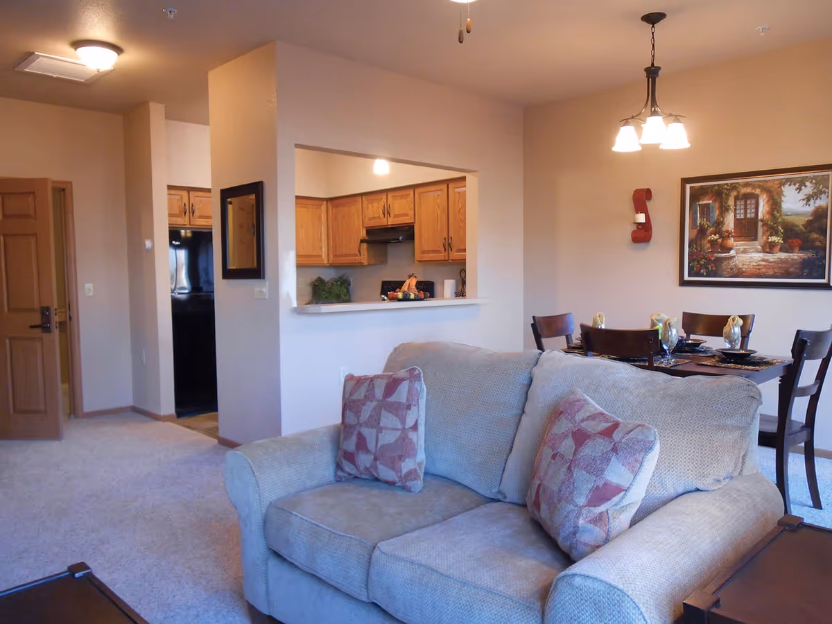 Interior view of a retirement community apartment showing a beige couch with patterned pillows in the foreground, a dining table set with four chairs and place settings in the background, and a kitchen area with wooden cabinets and a black refrigerator visible through a pass-through window.