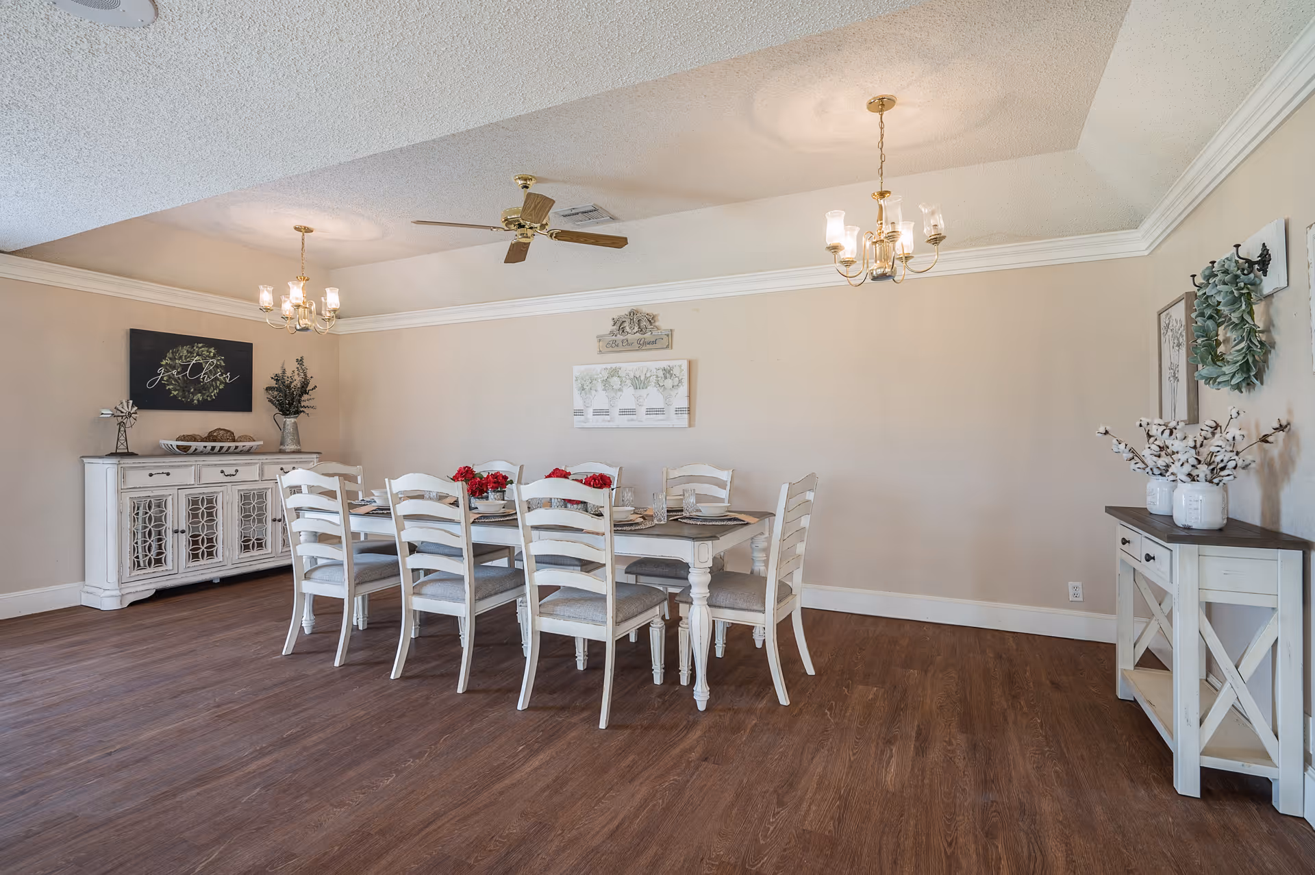 A spacious dining room with a long white dining table surrounded by eight matching chairs. The table is set with plates and decorated with red flowers. The room features wooden flooring, beige walls, two chandeliers, a ceiling fan, and decorative furniture including a sideboard and a console table with plants and artwork on the walls.