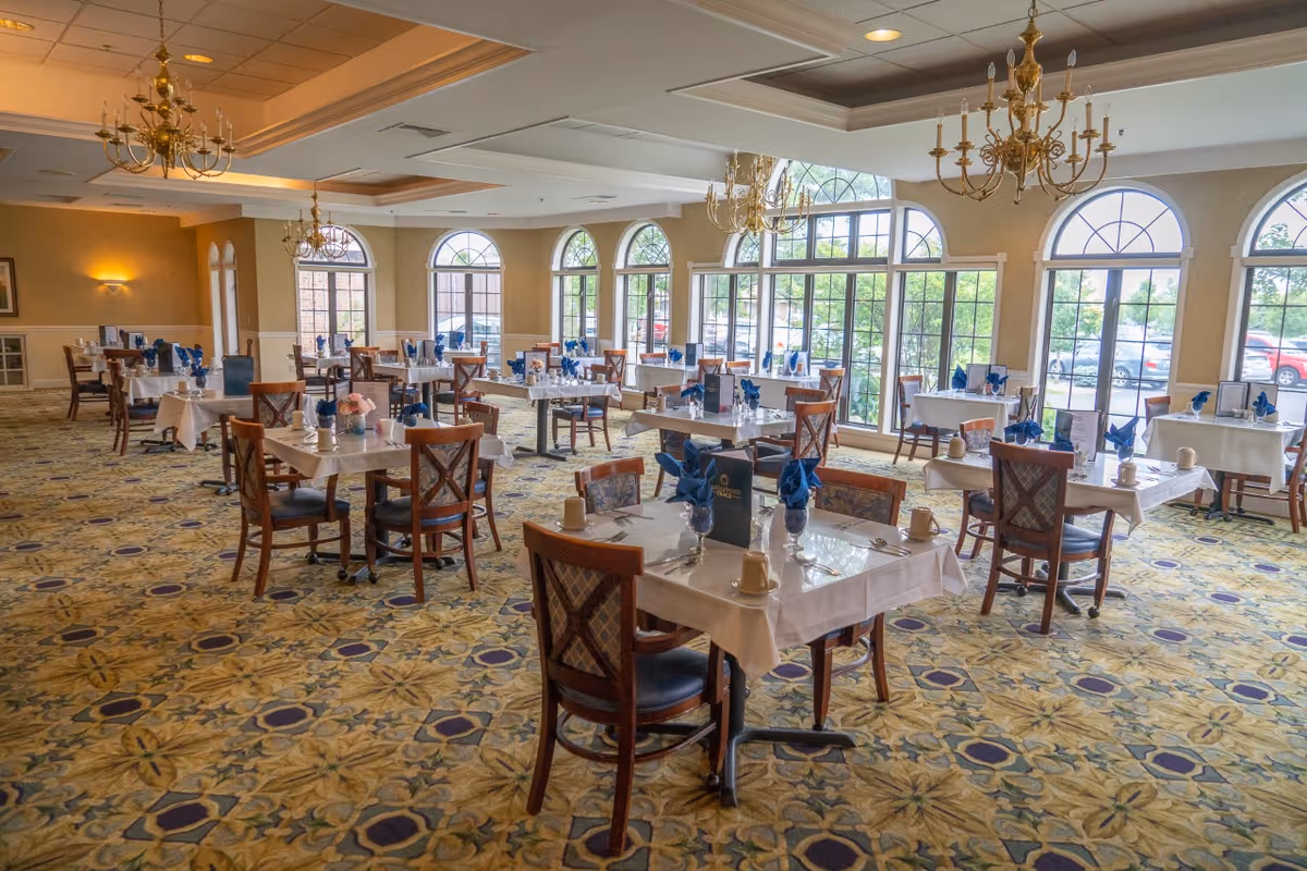 A spacious dining room in a senior living facility with multiple tables covered in white tablecloths, each set with blue napkins, glasses, and cups. The room features large arched windows allowing natural light to fill the space, patterned carpet flooring, and elegant chandeliers hanging from the ceiling.
