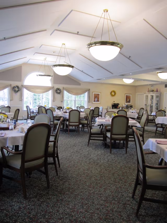 A spacious dining room with multiple round tables covered with white tablecloths and set with cups, glasses, and napkins. The room has large windows with beige curtains, decorative wreaths on the walls, framed pictures, and elegant hanging light fixtures. The carpet has a patterned design, and the chairs have wooden frames with light green upholstery.