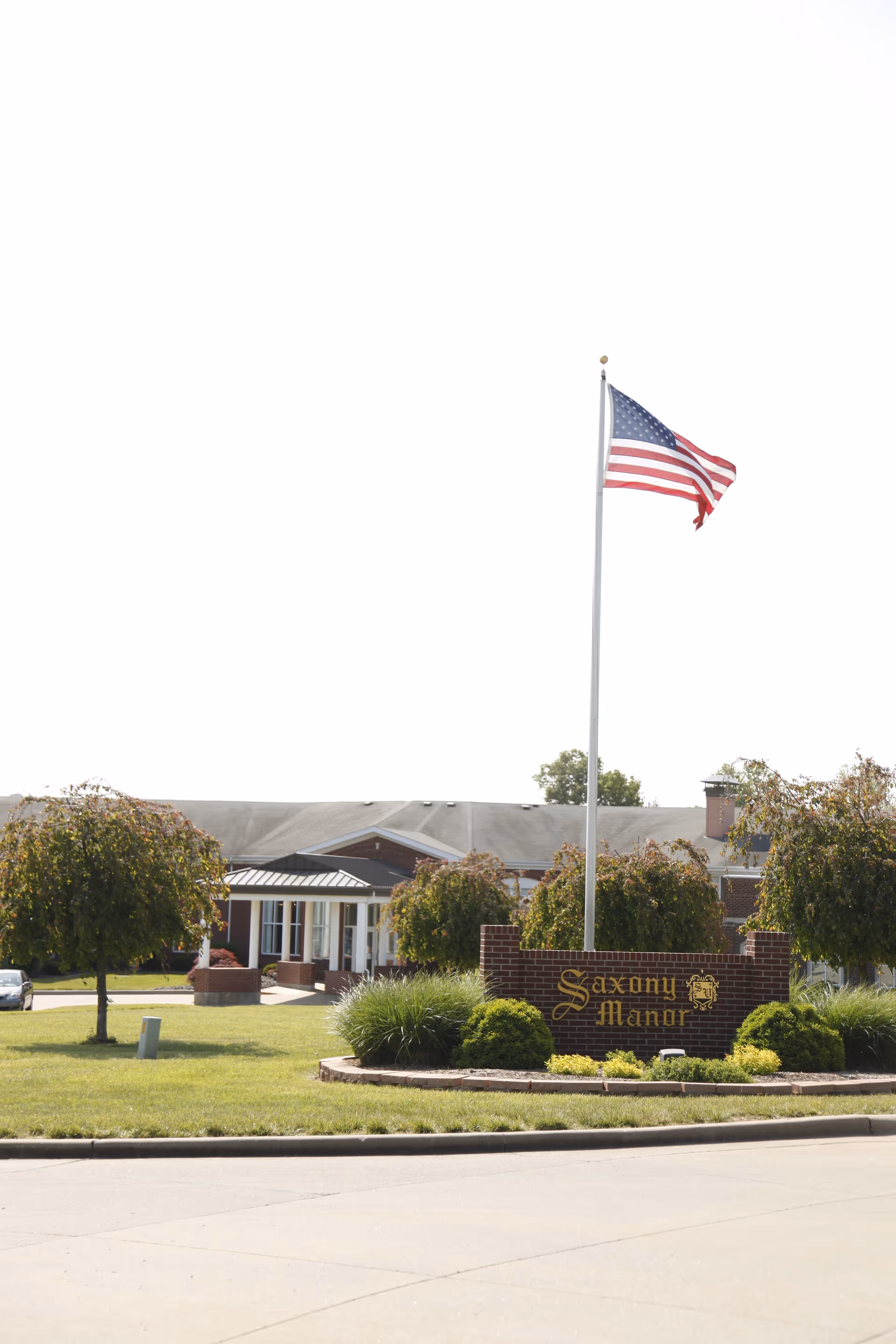 Exterior view of Saxony Manor, a brick building with a covered entrance, surrounded by green lawns and trees. In front of the building is a brick sign with the name 'Saxony Manor' and an American flag flying on a tall flagpole.