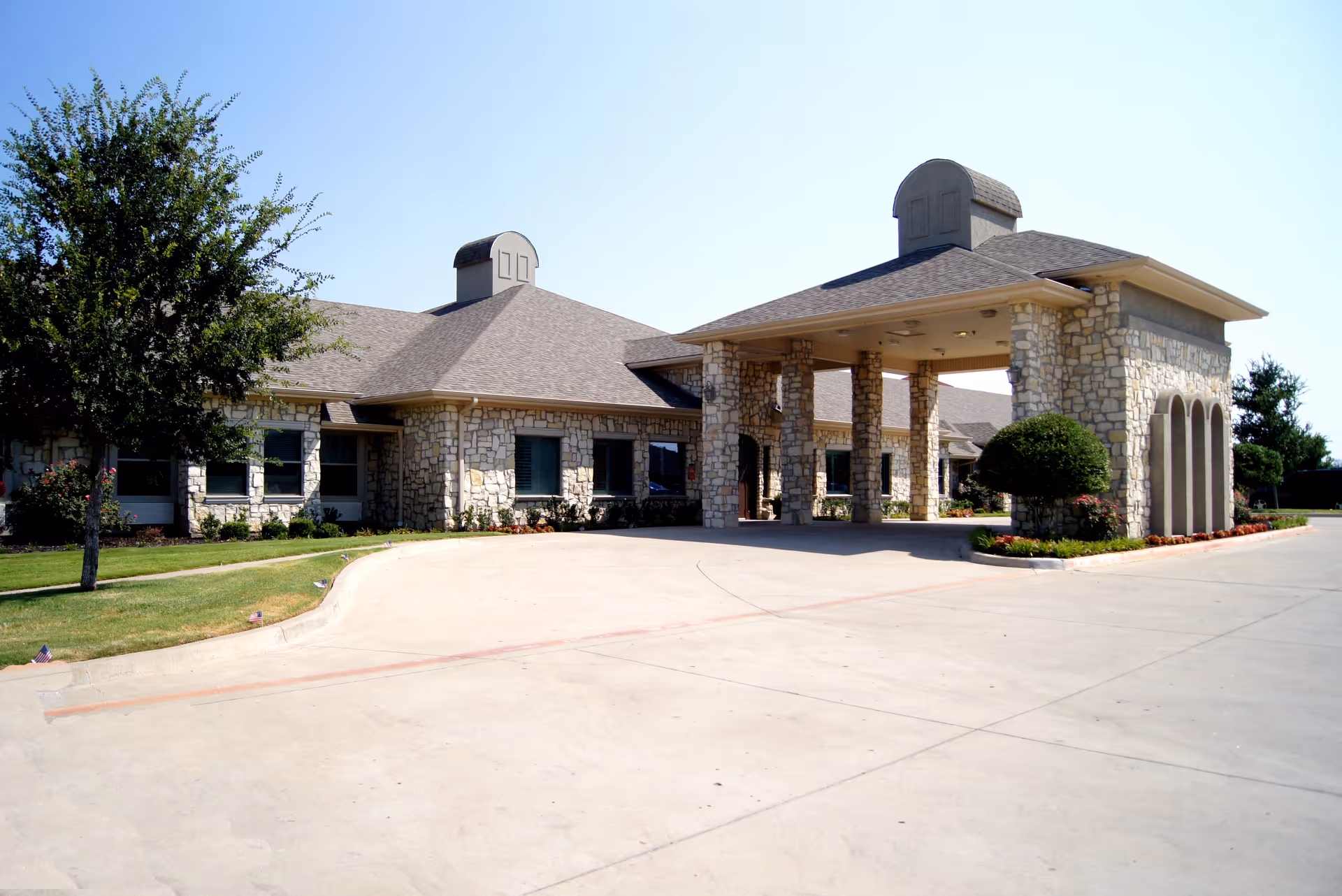 Exterior view of a single-story stone building with a covered entrance supported by stone pillars, surrounded by a paved driveway and landscaped greenery including trees and bushes under a clear blue sky.