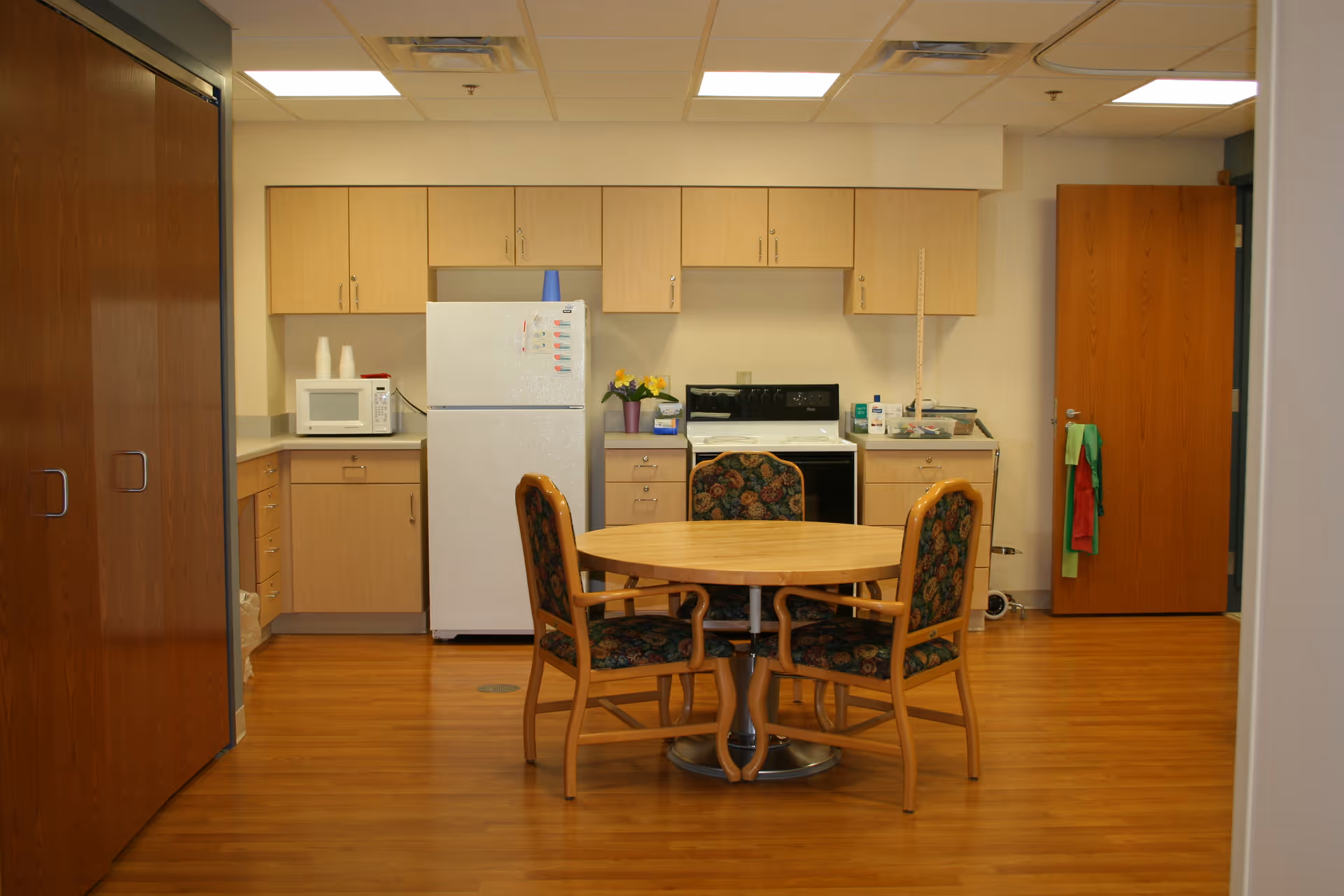 A small kitchen area with light wood cabinets, a white refrigerator, a microwave, and a stove. In front of the kitchen is a round wooden table with four chairs that have floral upholstery. The floor is wooden, and there are two closed wooden doors on either side of the kitchen.