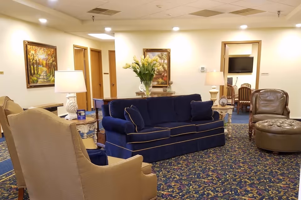 A cozy living room area in a senior living facility featuring a navy blue sofa with matching cushions, a beige armchair, a brown leather chair with a matching ottoman, two white table lamps on side tables, floral paintings on the walls, and a vase with yellow flowers on a wooden table. The room has patterned blue carpet and cream-colored walls with several doorways leading to other rooms.