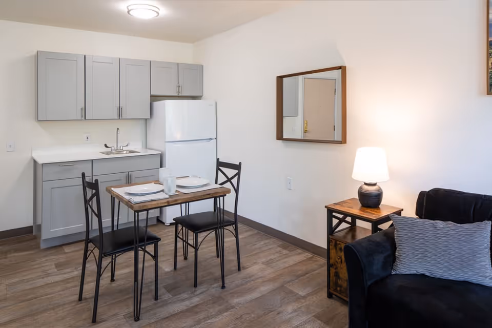 A small kitchen and dining area in a senior living facility with light gray cabinets, a white refrigerator, a small wooden table set for two with plates and mugs, a black cushioned chair, a black sofa with a gray pillow, a wooden side table with a lamp, and a wall-mounted mirror.