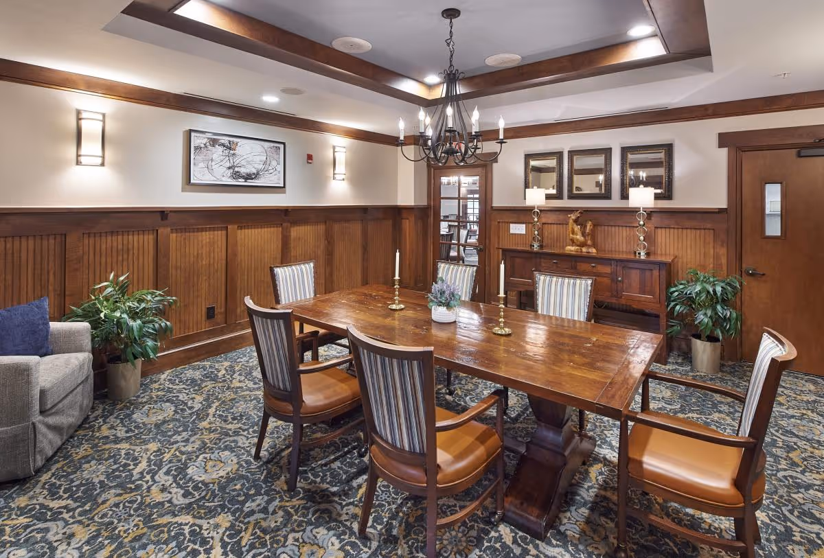A cozy dining room with a wooden rectangular table surrounded by six chairs with striped upholstery and leather seats. The room features wood paneling on the walls, a patterned carpet, two potted plants, a sideboard with decorative lamps and a sculpture, three mirrors above the sideboard, and a chandelier hanging from a tray ceiling with recessed lighting.