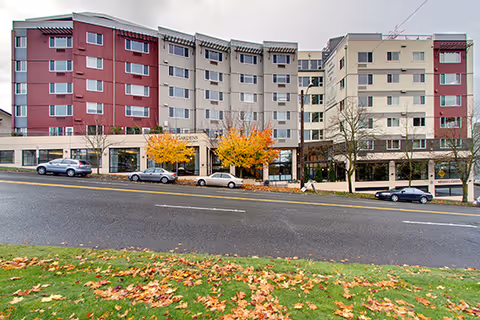 Exterior front facade of the Brookdale West Seattle senior living building along a street with parked cars and autumn trees on the sidewalk.