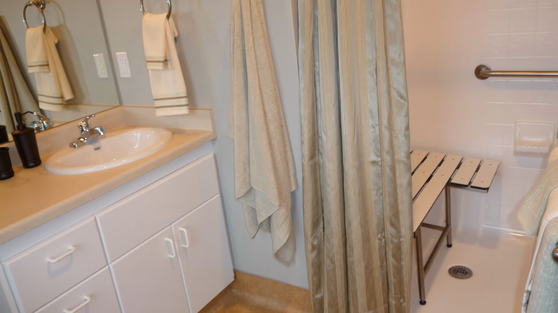 A bathroom with a white sink and countertop, white cabinets below, a large mirror above the sink, beige towels hanging on a ring and a hook, and a shower area with a beige striped curtain, a white shower bench, and a grab bar on the tiled wall.