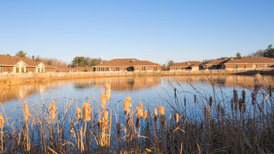 A pond with cattails in the foreground reflecting a row of single-story brick senior living buildings under a clear blue sky.
