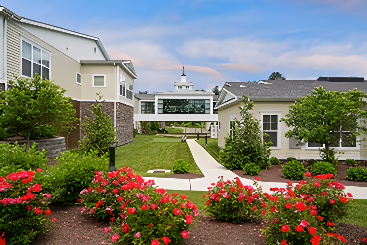 Outdoor view of Church Hill Village showing a well-maintained garden with red flowers and green shrubs in the foreground, a paved walkway leading between two buildings, and a skybridge connecting the buildings under a partly cloudy sky.
