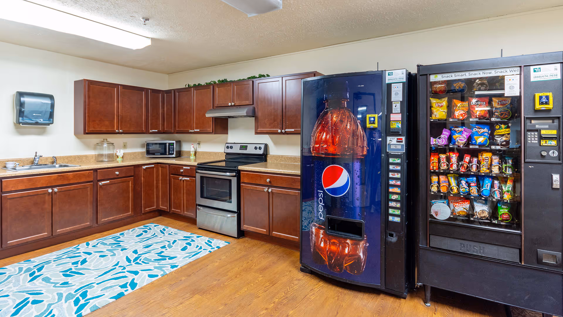 A communal kitchen area with wooden cabinets, a stainless steel electric stove, a microwave, and a sink. There is a blue and white patterned rug on the wooden floor. Two vending machines are present: one for Pepsi beverages and another stocked with various snacks.