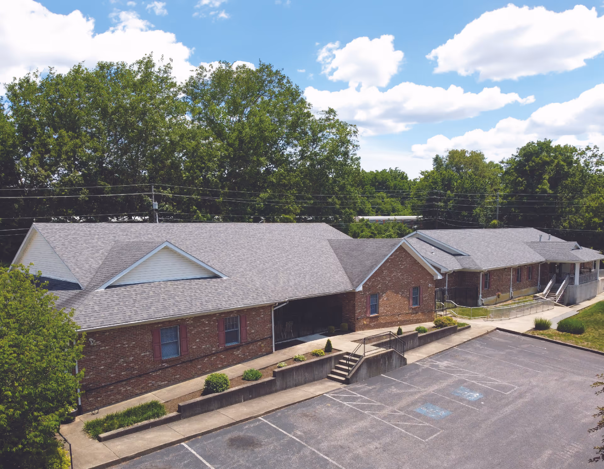 Exterior view of a single-story brick building with a gray shingled roof, surrounded by trees and a parking lot with marked spaces including handicapped spots, under a partly cloudy sky.