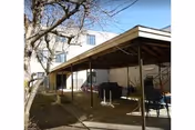 Outdoor covered patio area with metal roof and support poles adjacent to a multi-story building. There are some chairs and a grill under the patio, with a leafless tree nearby and a clear blue sky above.