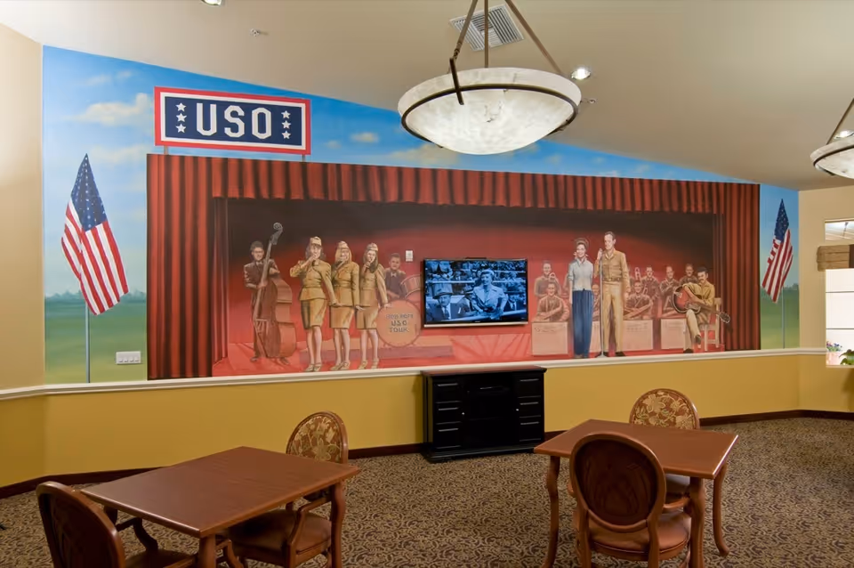 A common area with tables and chairs featuring a large mural on the wall depicting a USO stage show with performers in vintage military attire and American flags on either side. A flat-screen TV is mounted in the center of the mural showing a black and white image.