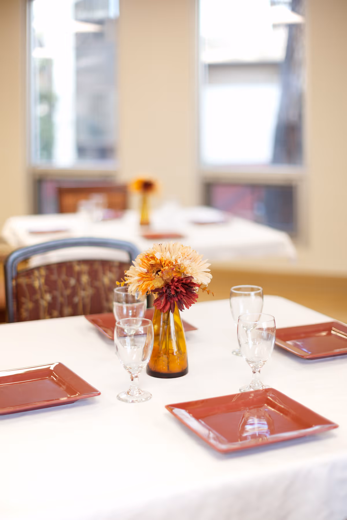 A dining table set with a white tablecloth, four red square plates, four clear water glasses, and a small amber vase with orange and red flowers in the center. In the background, there are windows and another similarly set table.