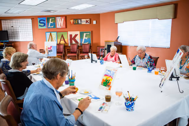 A group of elderly people seated around a large rectangular table in a brightly lit room, engaged in painting activities with watercolor paints and brushes. The walls are decorated with colorful letters spelling out 'SEW THINK RAKE'. Various art supplies and glasses with beverages are on the table.