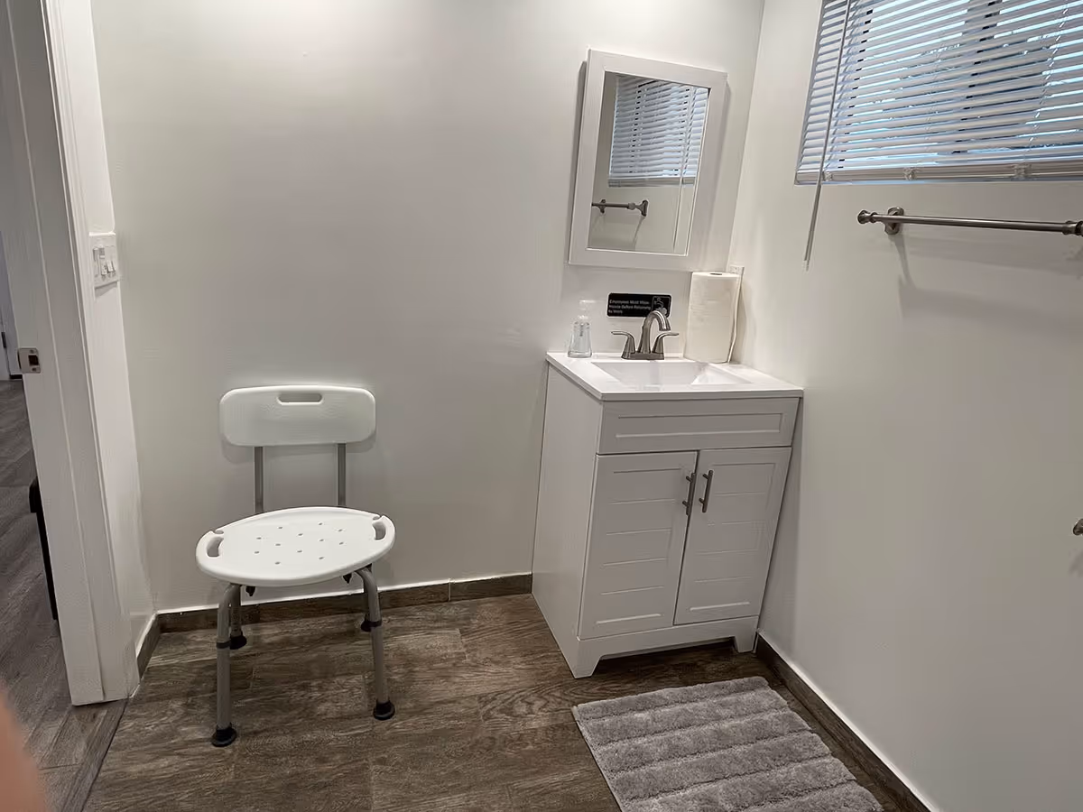 Small white bathroom interior featuring a plastic shower chair, a white sink vanity with mirror, towel bar, window blinds and a gray floor mat.