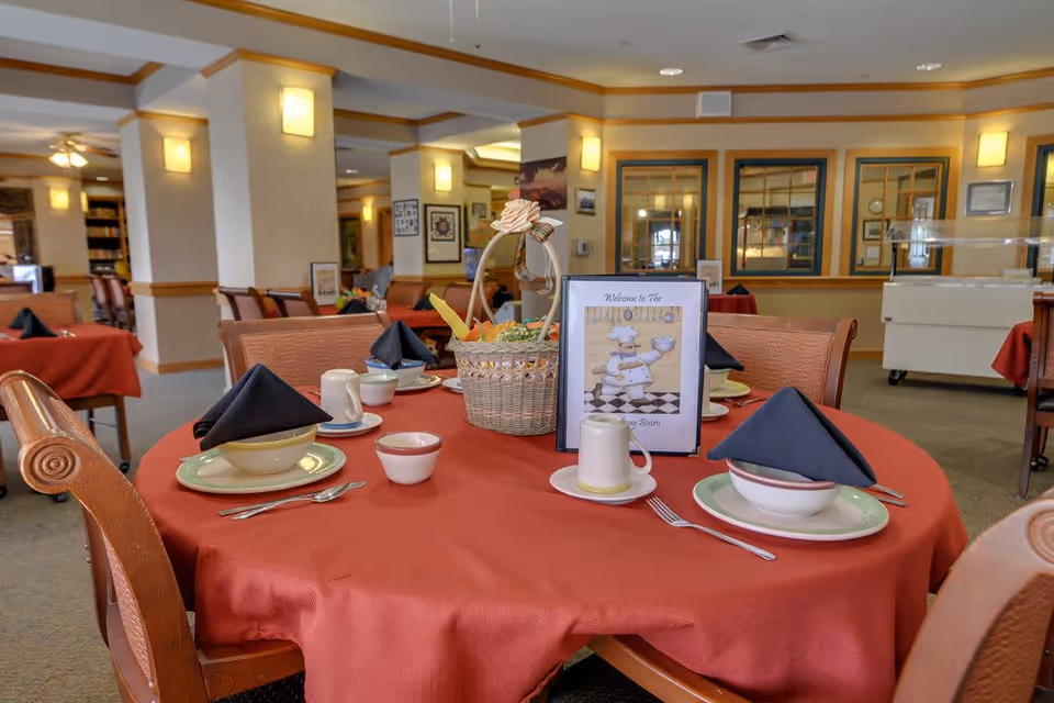 A dining room with a round table covered in a red tablecloth set for four people with plates, bowls, cups, utensils, and black folded napkins. A basket with decorative items and a menu card featuring a chef illustration are placed in the center of the table. The room has warm lighting, wooden chairs, and framed pictures on the walls.