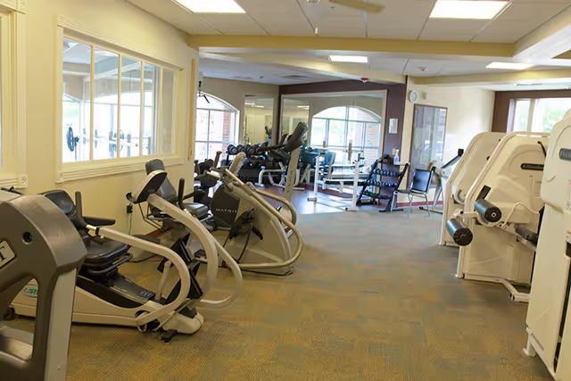 Interior view of a fitness room in a senior living facility with exercise equipment including stationary bikes, weight machines, and free weights. Large windows allow natural light to enter the room.