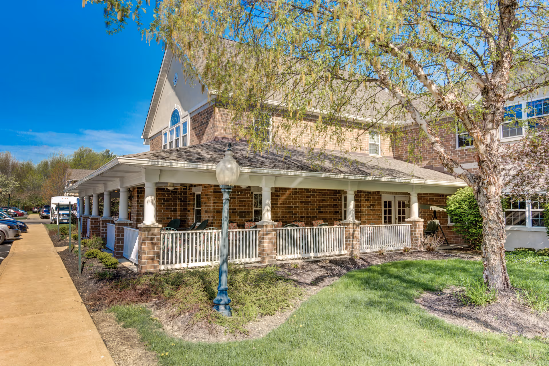 Exterior view of a brick building with a covered porch featuring white railings and columns. There is a sidewalk and parked cars to the left, a lamp post in front of the porch, and a tree with green leaves on the right side. The sky is clear and blue.