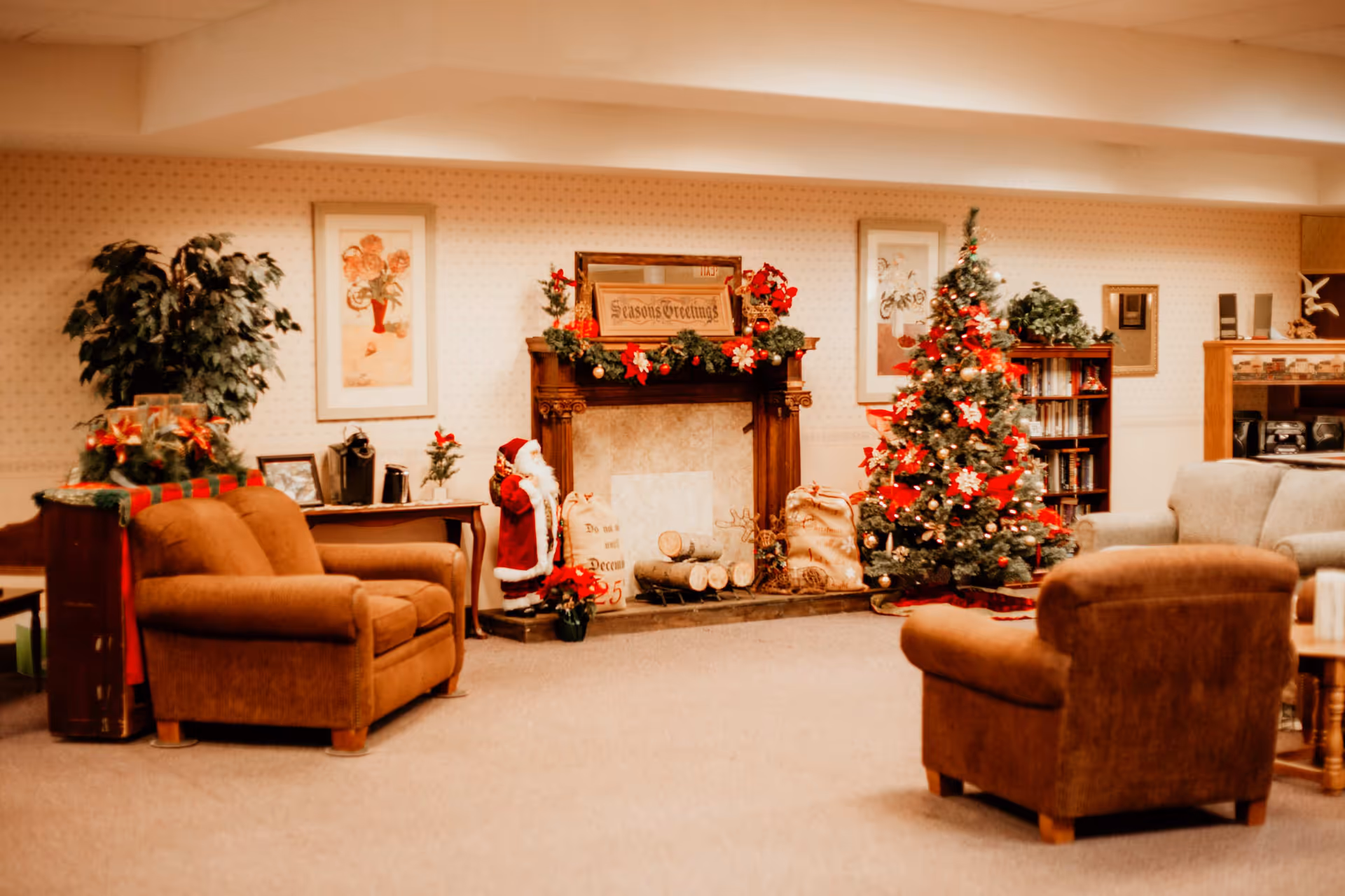 A cozy senior living community lounge decorated for Christmas with a Christmas tree adorned with red bows and ornaments, a fireplace mantel decorated with garlands and a 'Seasons Greetings' sign, a Santa Claus figure, and comfortable brown armchairs arranged around the room.