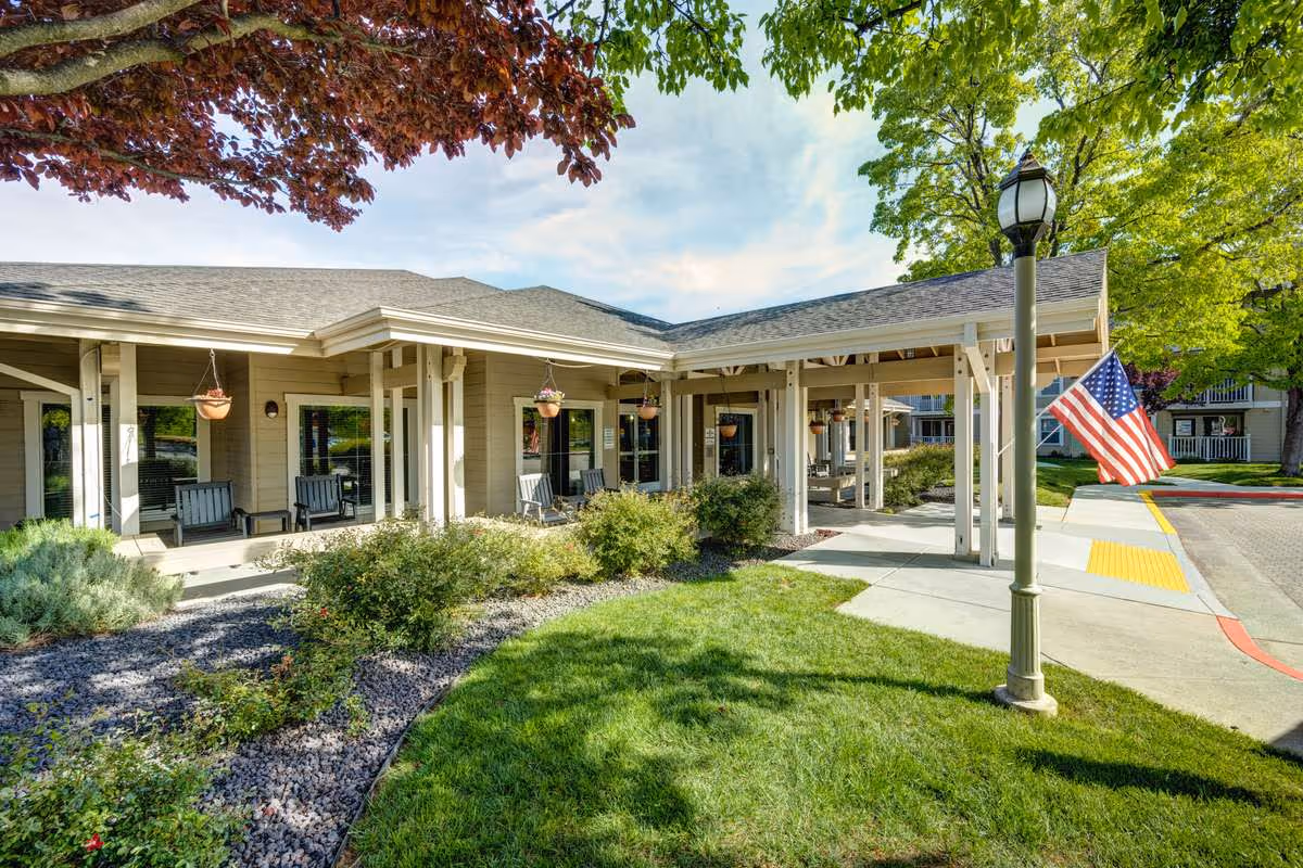 Exterior view of a senior living facility with a covered porch area featuring several chairs and hanging flower pots. The building is surrounded by well-maintained landscaping, including green grass, bushes, and trees. An American flag is displayed near a lamp post along a paved walkway.