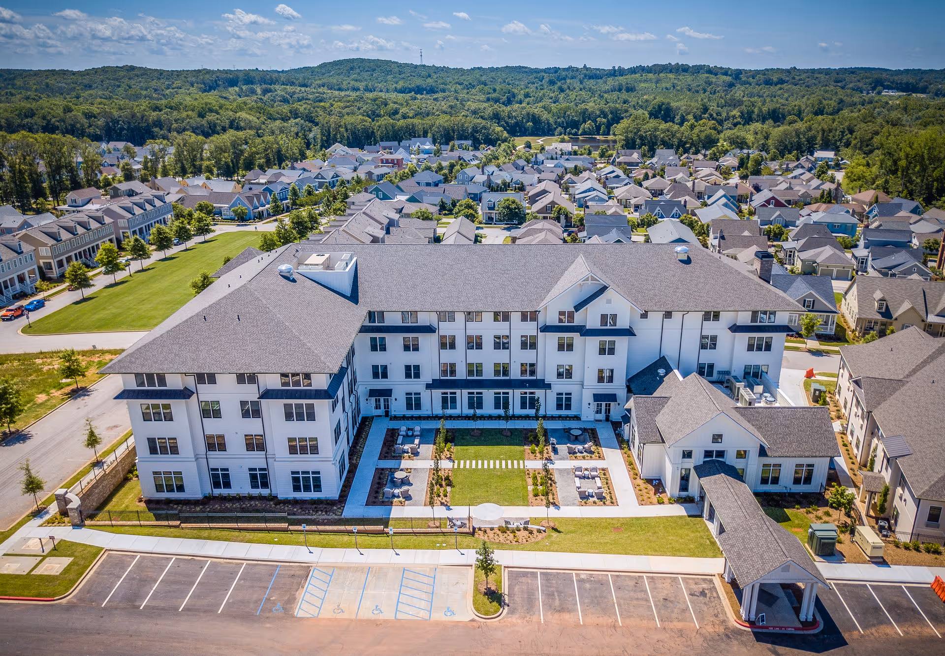 Aerial view of a three-story senior living building with a central landscaped courtyard, parking area, and surrounding neighborhood.