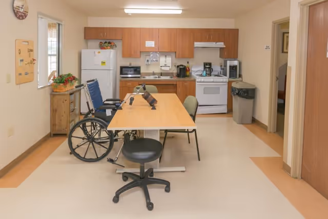 Communal kitchen and dining area with a table, chairs and a wheelchair in front of a kitchenette with refrigerator and stove.