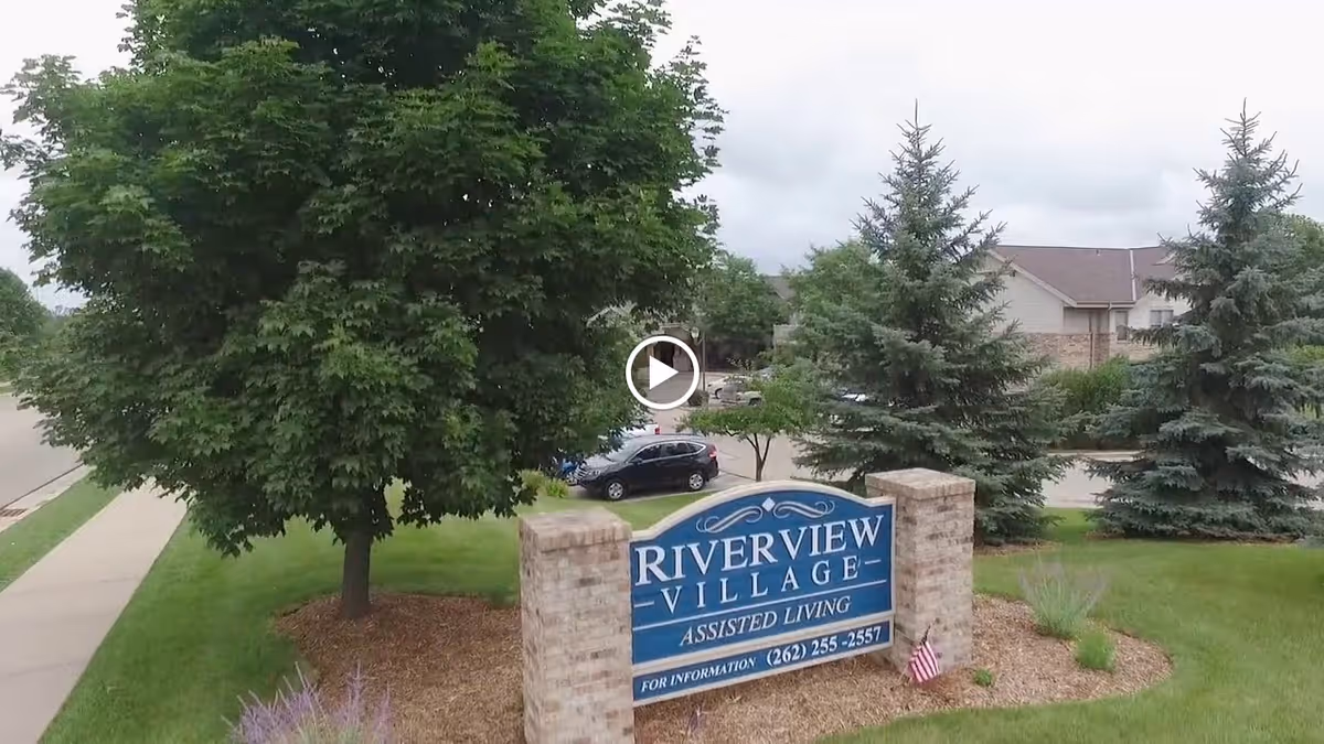 Outdoor view of the entrance sign for Riverview Village Assisted Living, surrounded by trees, grass, and landscaping with a sidewalk and parked cars in the background under a cloudy sky.