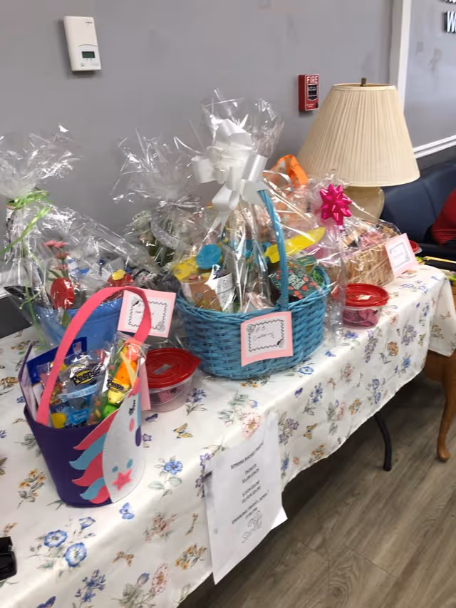 A table covered with a floral tablecloth displaying several gift baskets wrapped in clear plastic with bows. The baskets contain various items and are labeled with small cards. A lamp with a beige shade is on the right side of the table, and a gray wall with a thermostat and fire alarm is in the background.