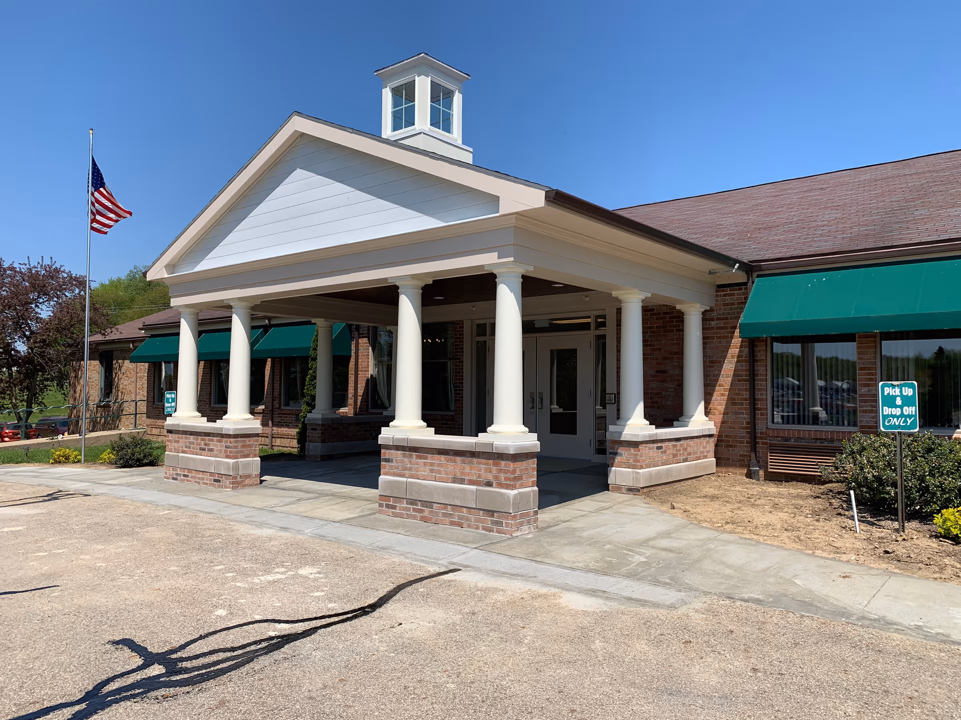 Front exterior view of Country Court Skilled Nursing Center showing a covered entrance with white columns and brick bases, green awnings over windows, an American flag on a flagpole, and a sign indicating pick up and drop off only.