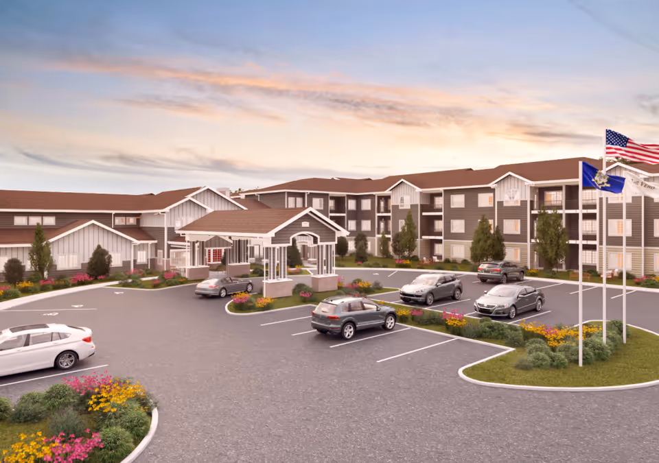 Exterior view of a senior living facility with a large parking lot, landscaped flower beds, and three flagpoles displaying the American flag and other flags. The building is three stories tall with a covered entrance and multiple windows, set against a sunset sky.
