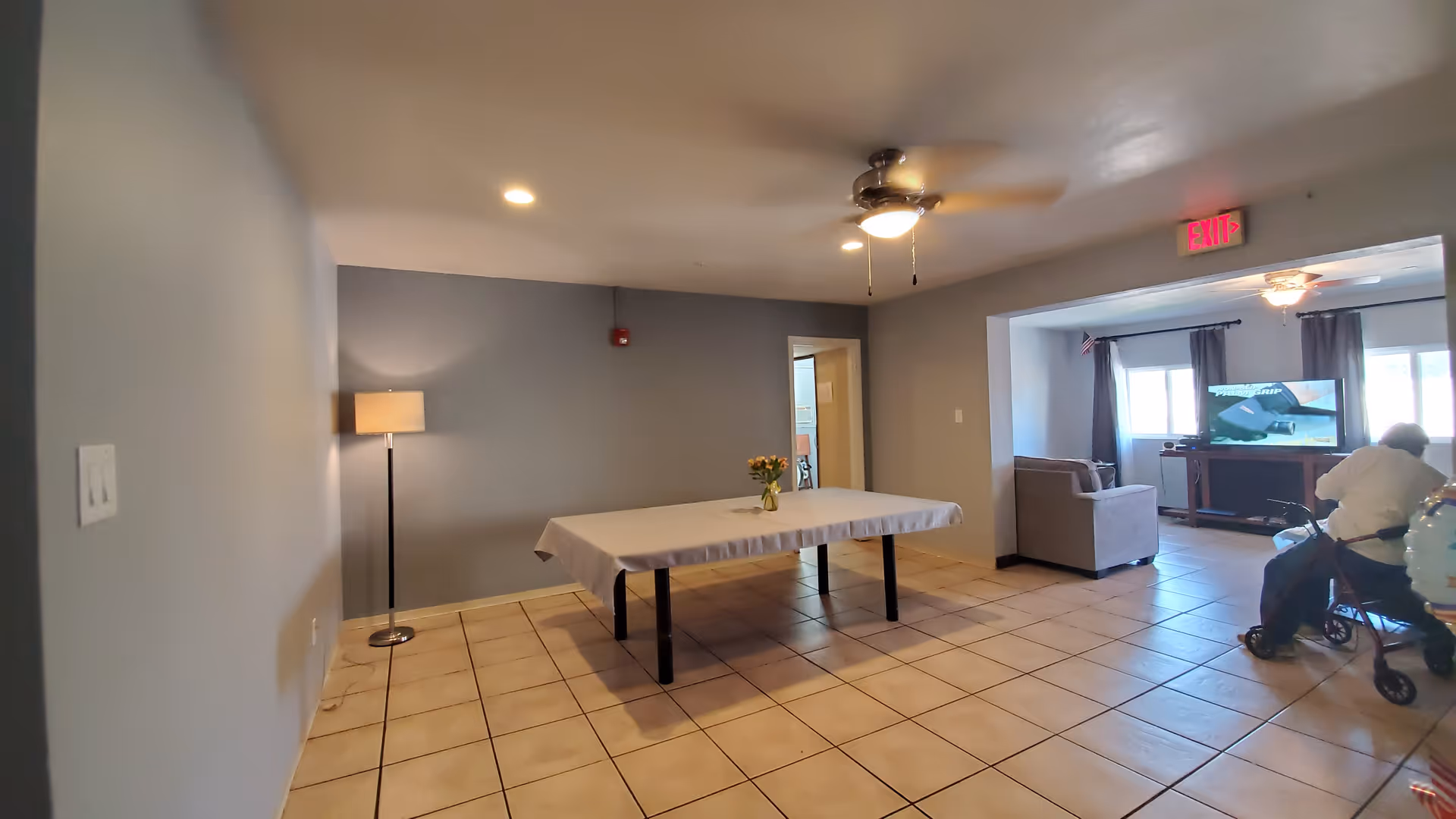 A spacious room with tiled floor and gray walls featuring a rectangular table covered with a white tablecloth and a small vase of flowers in the center. A floor lamp stands against the left wall. In the adjoining room, there is a person with a walker watching a television, with a couch and windows with curtains visible. A ceiling fan with a light is on the ceiling, and an exit sign is above the doorway.