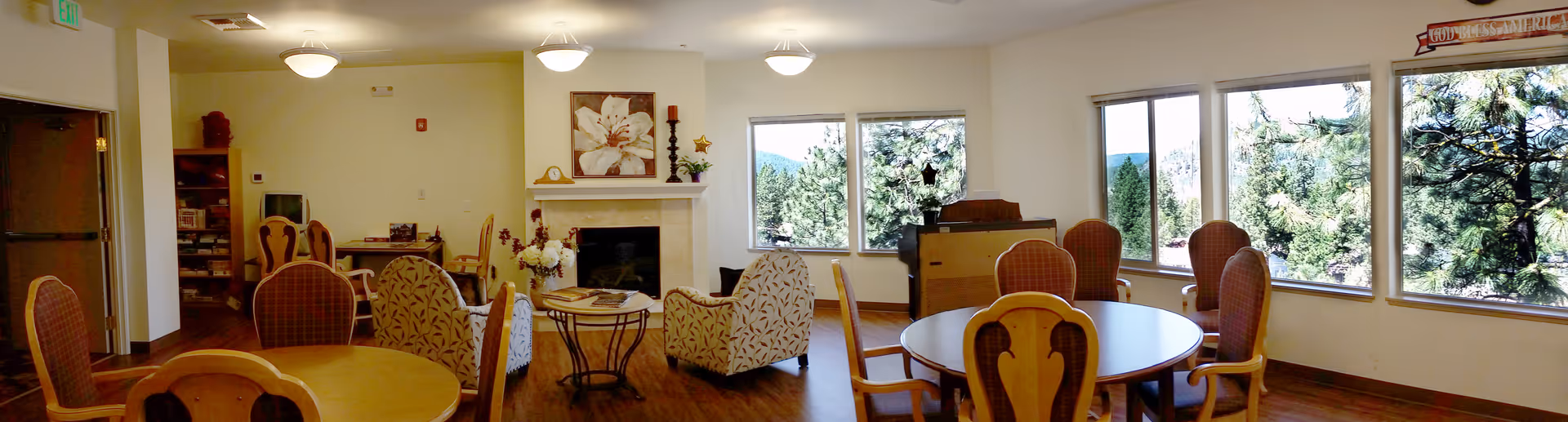 A spacious common area in a senior living facility with multiple round wooden tables and chairs arranged around the room. There are large windows on one side letting in natural light and showing a view of trees outside. The room features a fireplace with decorative items on the mantel, comfortable armchairs, and a piano near the windows. The flooring is wooden and the walls are painted light beige.