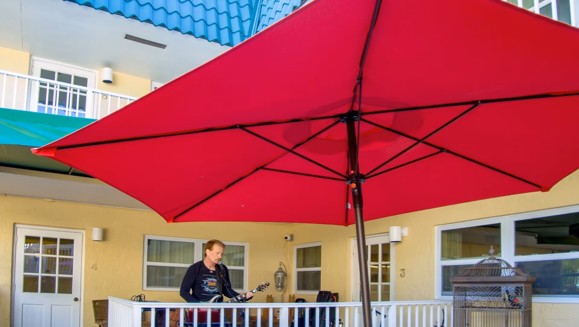 A man playing an electric guitar and singing into a microphone on a balcony or patio area with a large red umbrella overhead. The background shows yellow walls, white doors, and windows of a building.