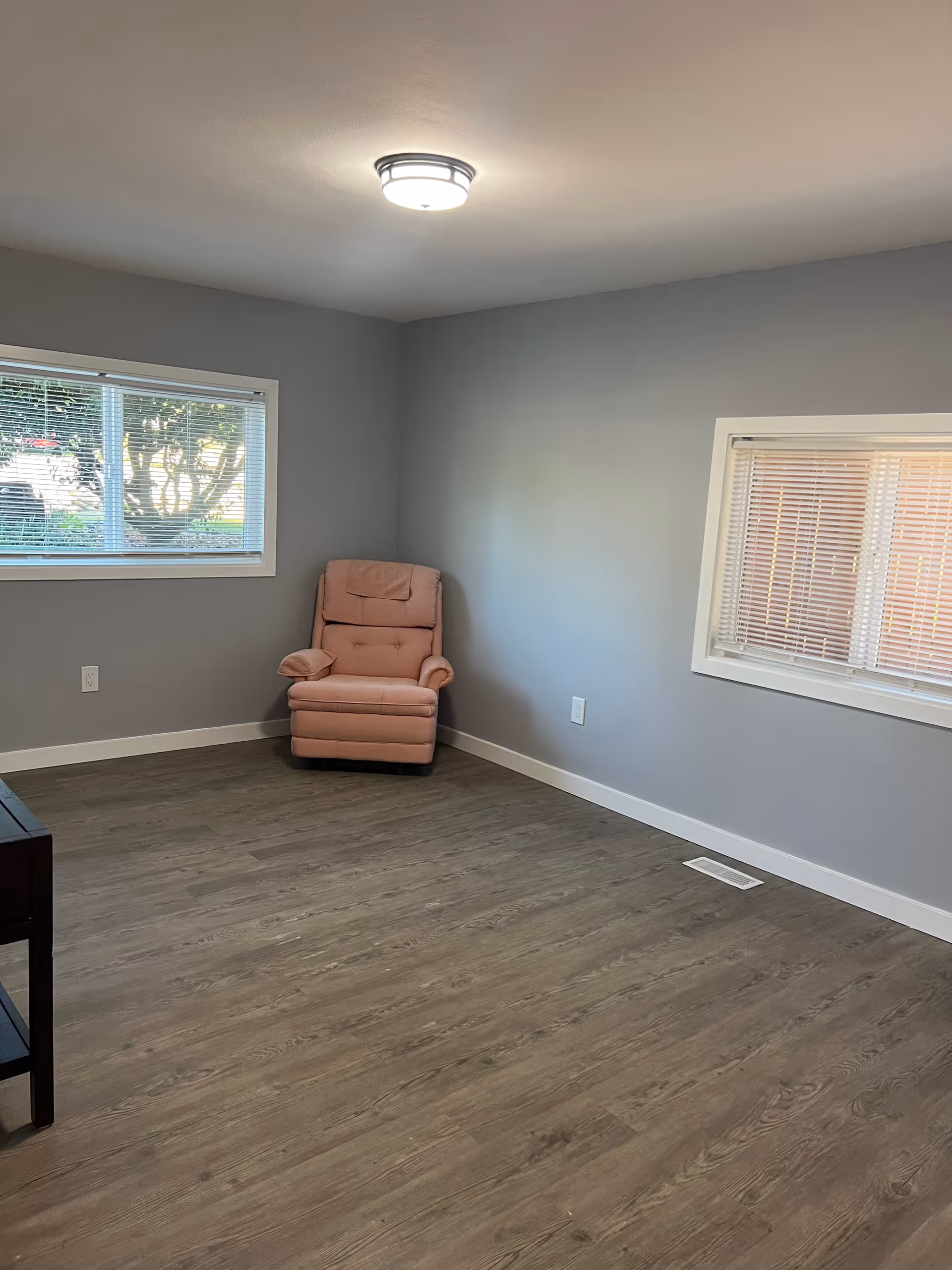 A sparsely furnished room with light gray walls and wood-look flooring. There is a single pink recliner chair positioned in the corner between two windows with blinds. A ceiling light fixture is mounted in the center of the ceiling.