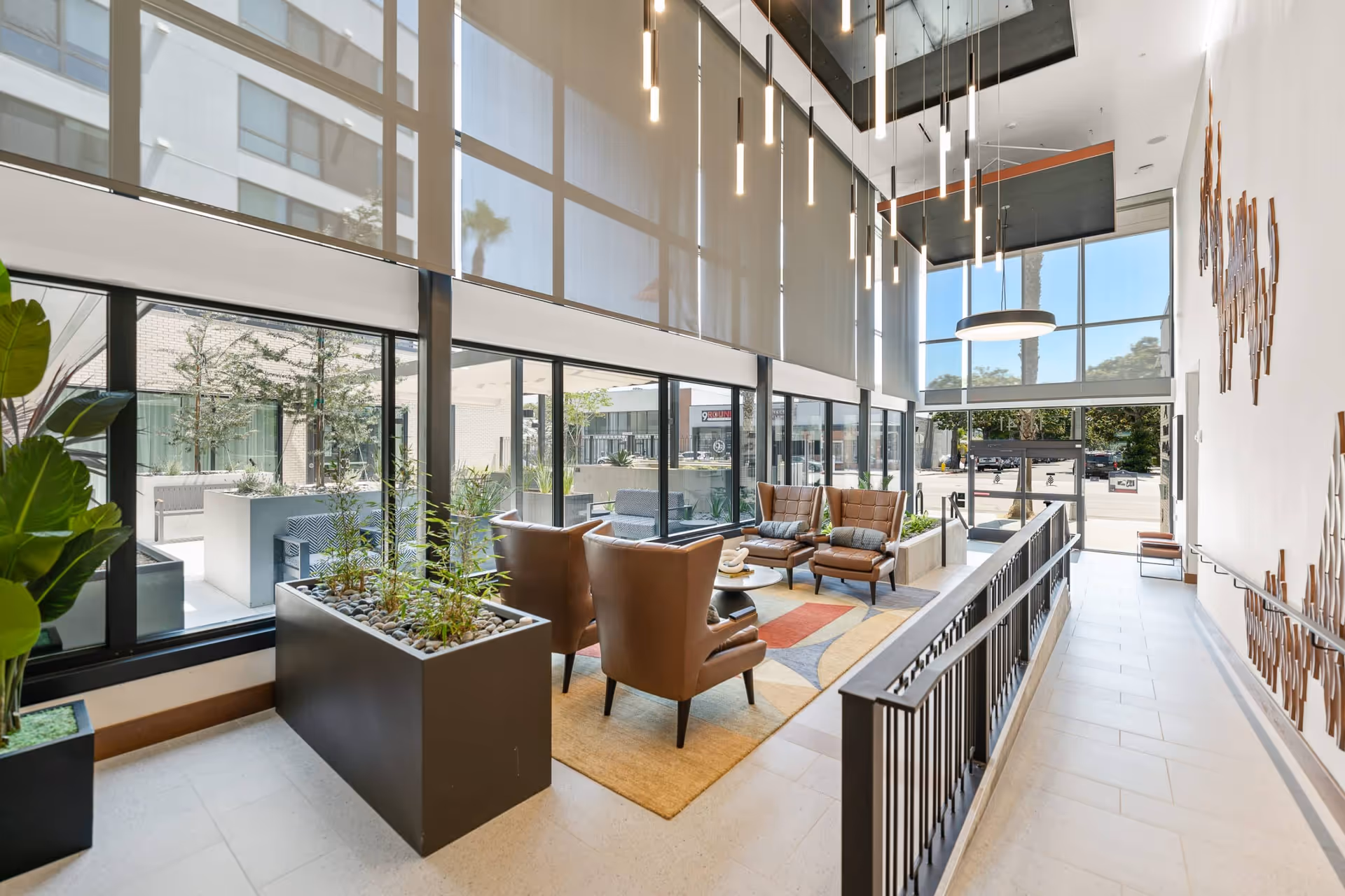 Modern interior lounge area with high ceilings and large windows. The space features four brown leather armchairs arranged around a circular coffee table on a colorful geometric rug. There are tall planters with green plants, hanging pendant lights, and a decorative wall installation. The area is adjacent to a glass door entrance with a view of the outdoor parking lot.