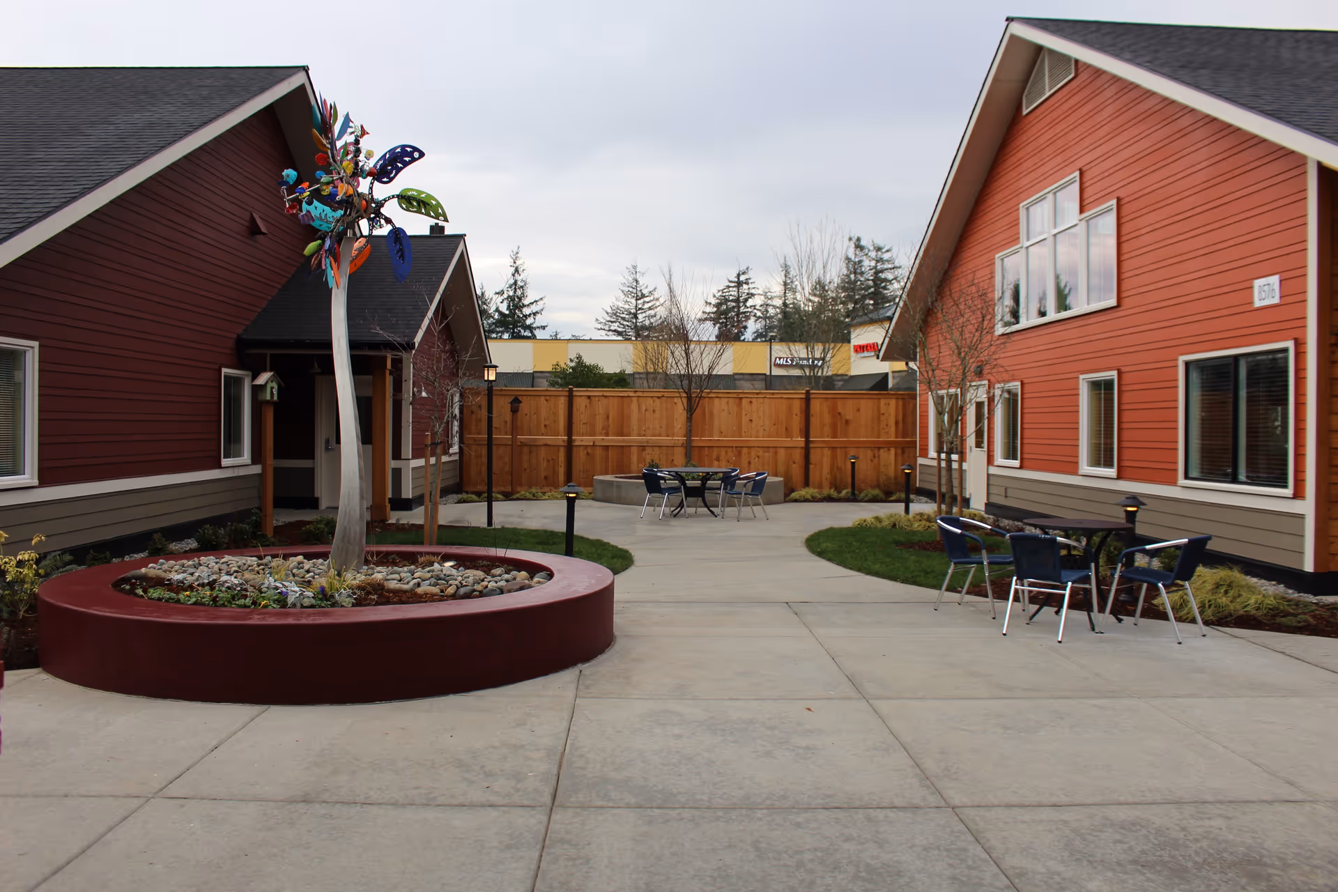 Outdoor courtyard area between two red buildings with gray roofs, featuring a circular planter with a colorful metal sculpture, several small trees, and patio tables with chairs on a concrete pathway.
