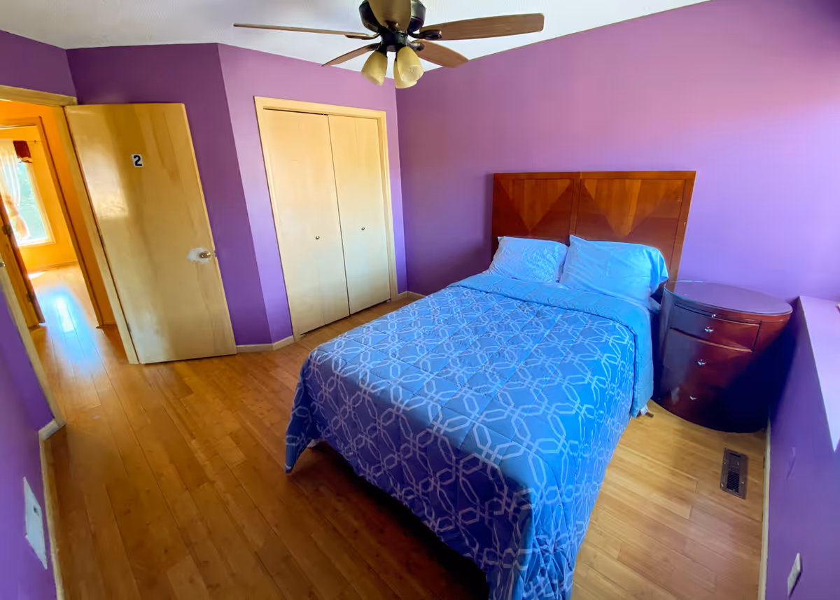 A bedroom with purple walls, a double bed with a patterned blue comforter, wooden headboard and nightstand, ceiling fan, closet, and hardwood floor.