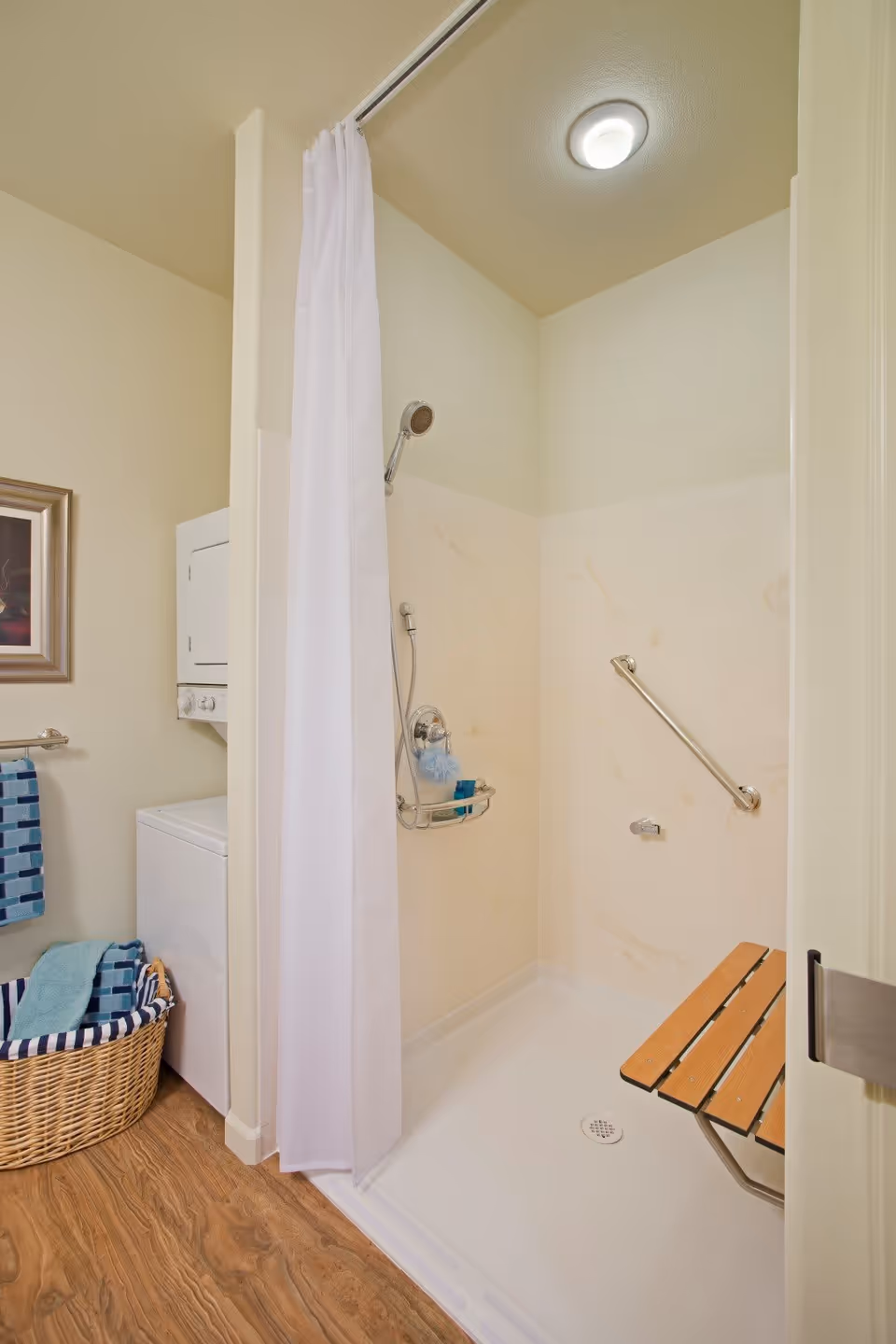 A bathroom shower area with a white shower curtain, a handheld showerhead, a grab bar on the wall, and a foldable wooden shower seat. Next to the shower is a stacked washer and dryer unit with a wicker basket containing folded towels on the floor. The walls are light-colored and the floor has a wood-like finish.
