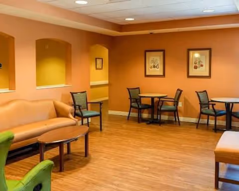 A cozy senior living common area with wooden flooring, orange walls, and recessed ceiling lights. The room features a tan leather sofa, several wooden chairs with cushions, round tables, and framed artwork on the walls.