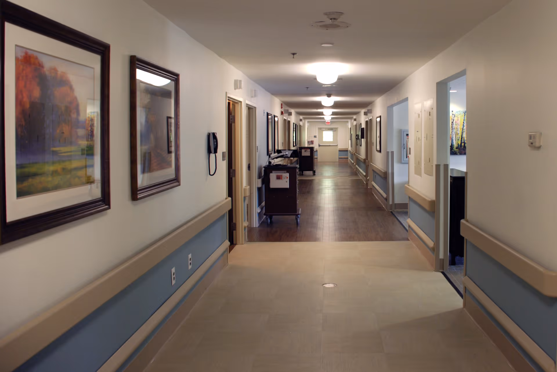 Long interior hallway of a senior living facility with framed artwork, handrails along the walls, ceiling lights, and service carts at the far end.